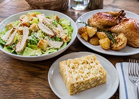 un plato de ensalada cesar, plato pollo al horno y plato brownie de chocolate blanco en una mesa